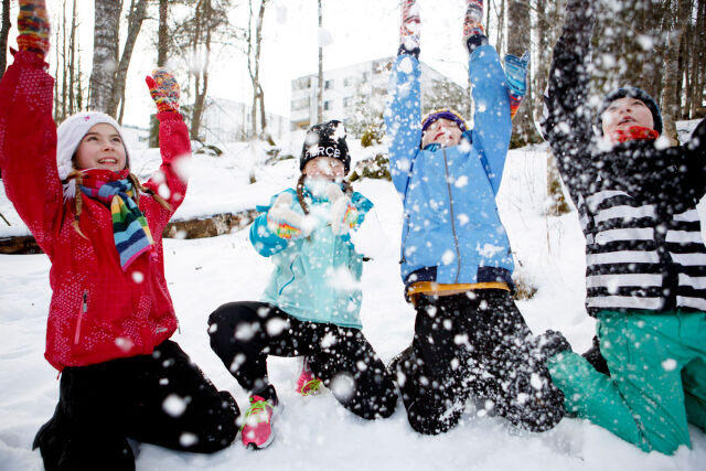 Neljä värikkäisiin talvivaatteisiin pukeutunutta lasta istuu polvillaan maassa ja heittää lunta ilmaan.