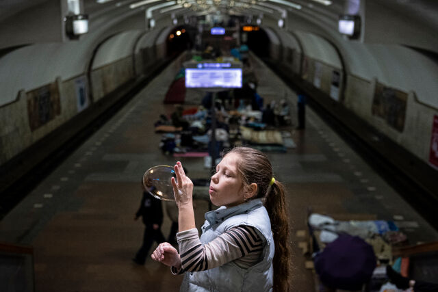 Tyttö puhaltaa suurta saippuakuplaa metroaseman ylätasanteella. Alhaalla asemalaiturilla on muita ihmisiä.