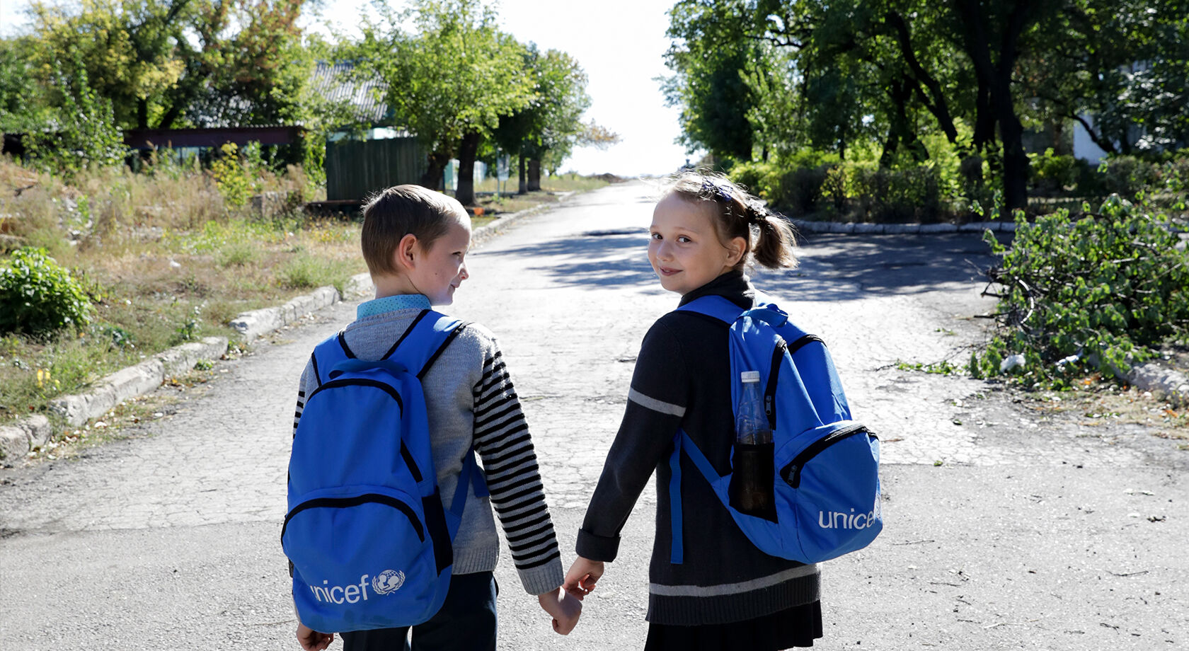 Tyttö ja poika kävelevät kouluun UNICEF-reput selässään ja pitävät toisiaan kädestä kiinni. Tyttö katsoo kameraan ja poika tyttöön hymyillen.