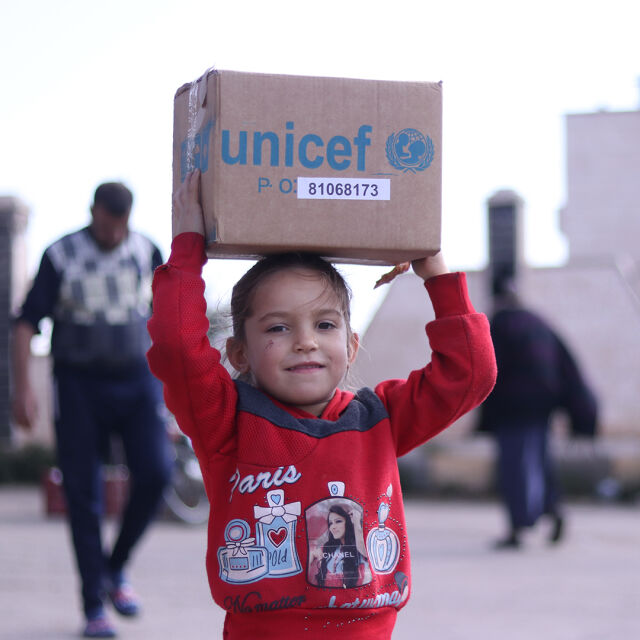 Iloinen lapsi kantaa UNICEFin vaaatelaatikkoa päänsä päällä.